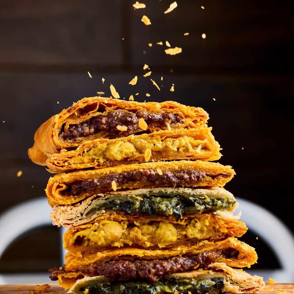 Stack of flaky golden pastries filled with savory meat and vegetables on a wooden board, with crumbs scattered around.
