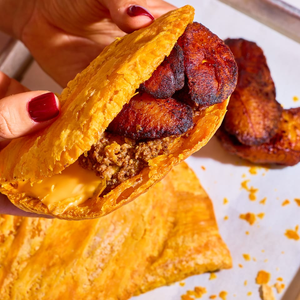 Hand holding a golden flaky pastry stuffed with seasoned meat, grilled sausage, and melted cheese over a tray of pastries.