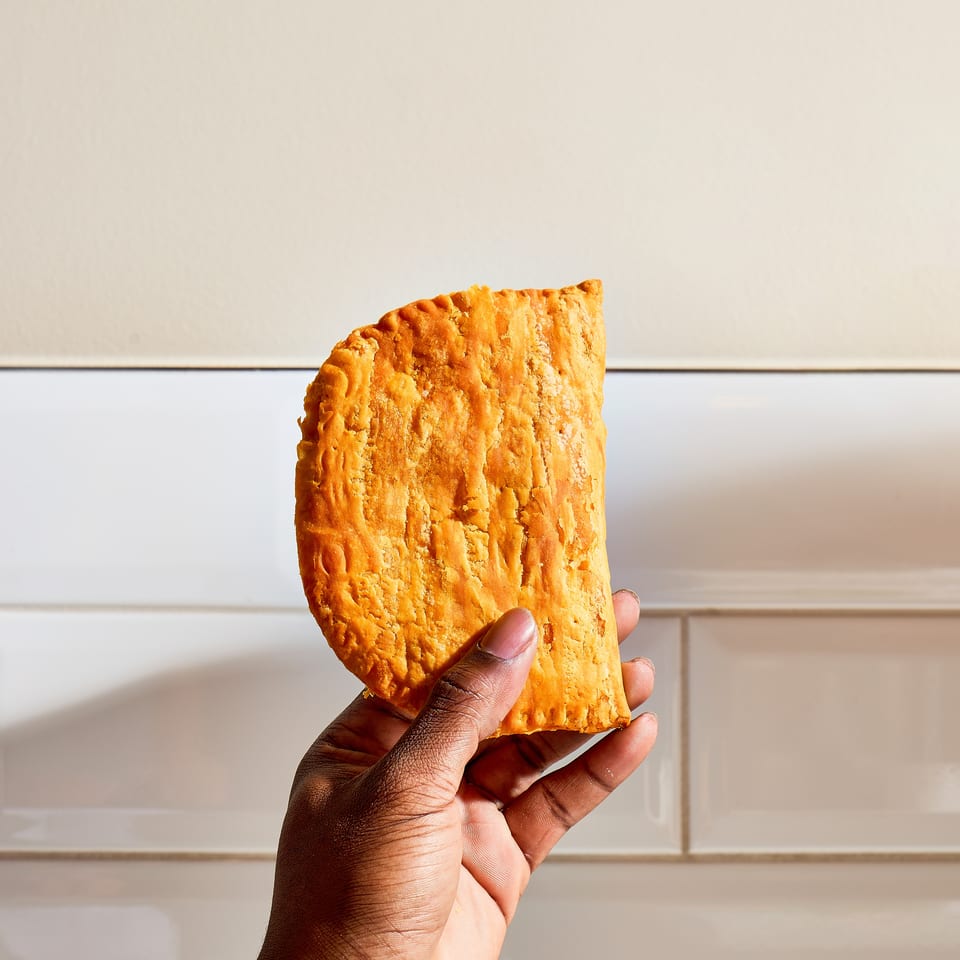 A golden, flaky folded pastry is held in one hand against a light tiled background.