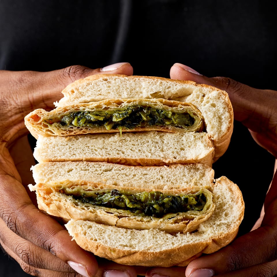 Hands hold a large sandwich layered with flaky pastry and dark green leafy filling against a black background.
