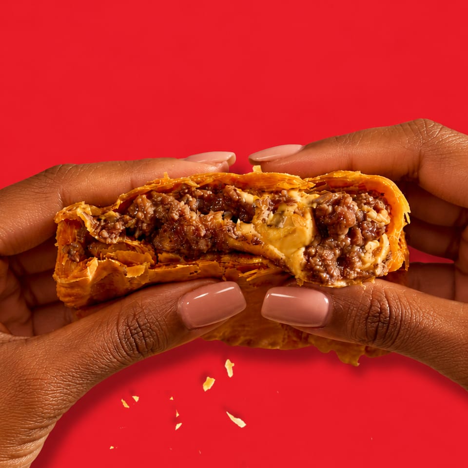 Hands with manicured nails hold a flaky pastry filled with seasoned ground meat and melted cheese against a bright red background.
