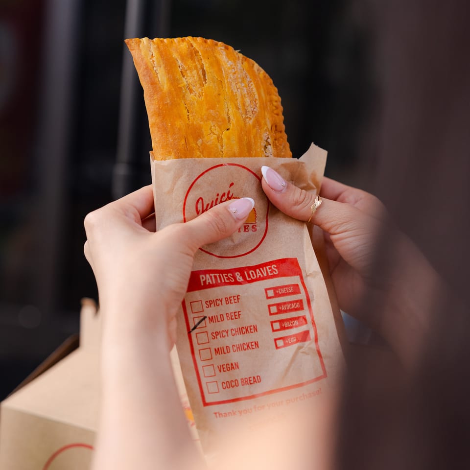 Hands hold a golden, flaky pastry in a branded paper bag against a dark background.