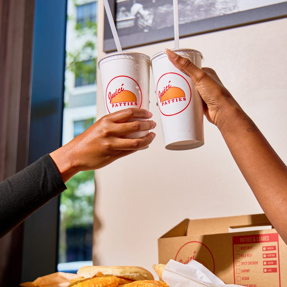 Two hands hold Juici Patties drinks above a counter displaying golden patties and branded takeaway packaging.