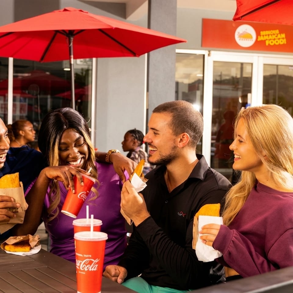Happy friends enjoy Jamaican food and drinks at an outdoor restaurant under red umbrellas.