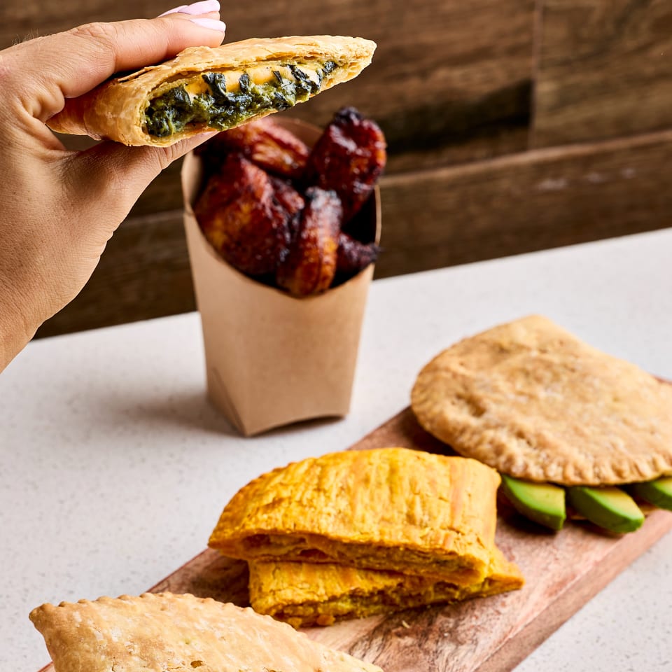 Hand holds a flaky spinach-filled pastry above a cup of fried snacks, with assorted stuffed pastries arranged on a wooden board.