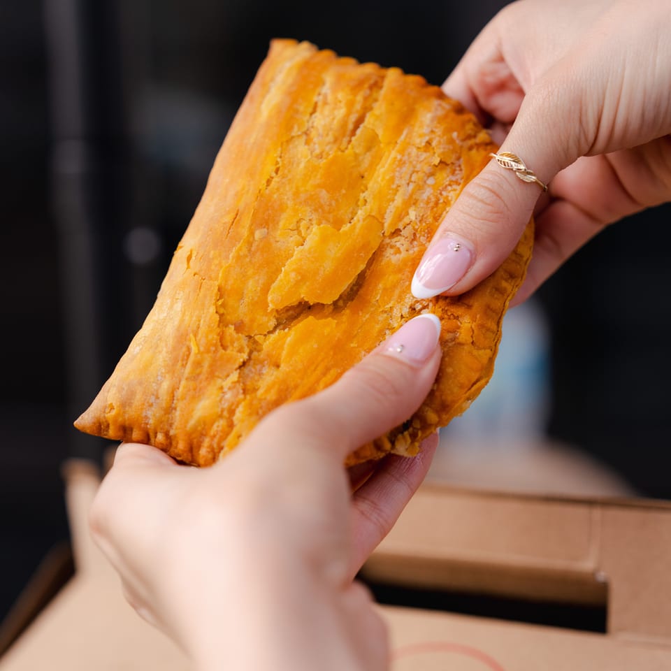 Hands hold a golden, flaky stuffed pastry over an open cardboard takeaway box.