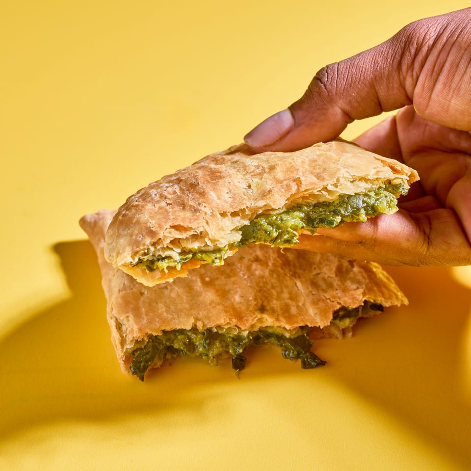 Hands hold a flaky stuffed pastry filled with green herbs against a bright yellow background.