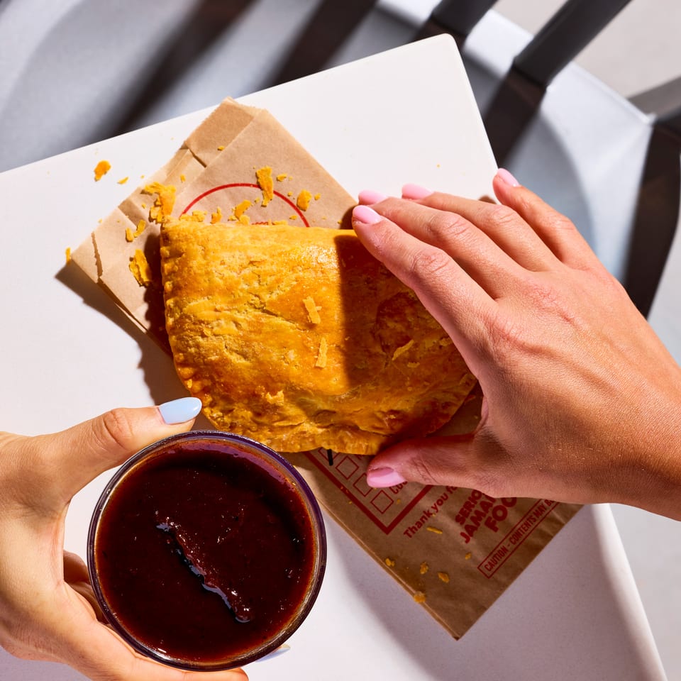 Hands share a golden flaky pastry on branded paper beside a cup of dark dipping sauce at an outdoor table.