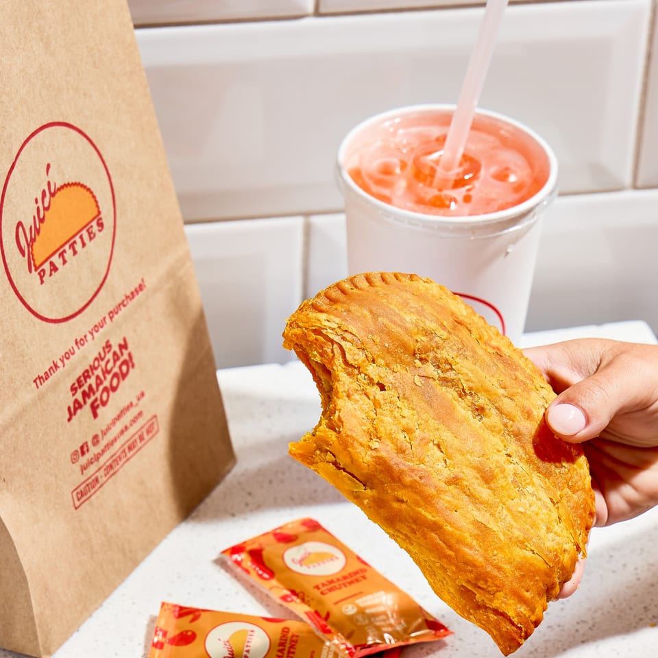 Hand holds a golden flaky sandwich beside a pink drink, branded paper bag, and packets of tamarind chutney.