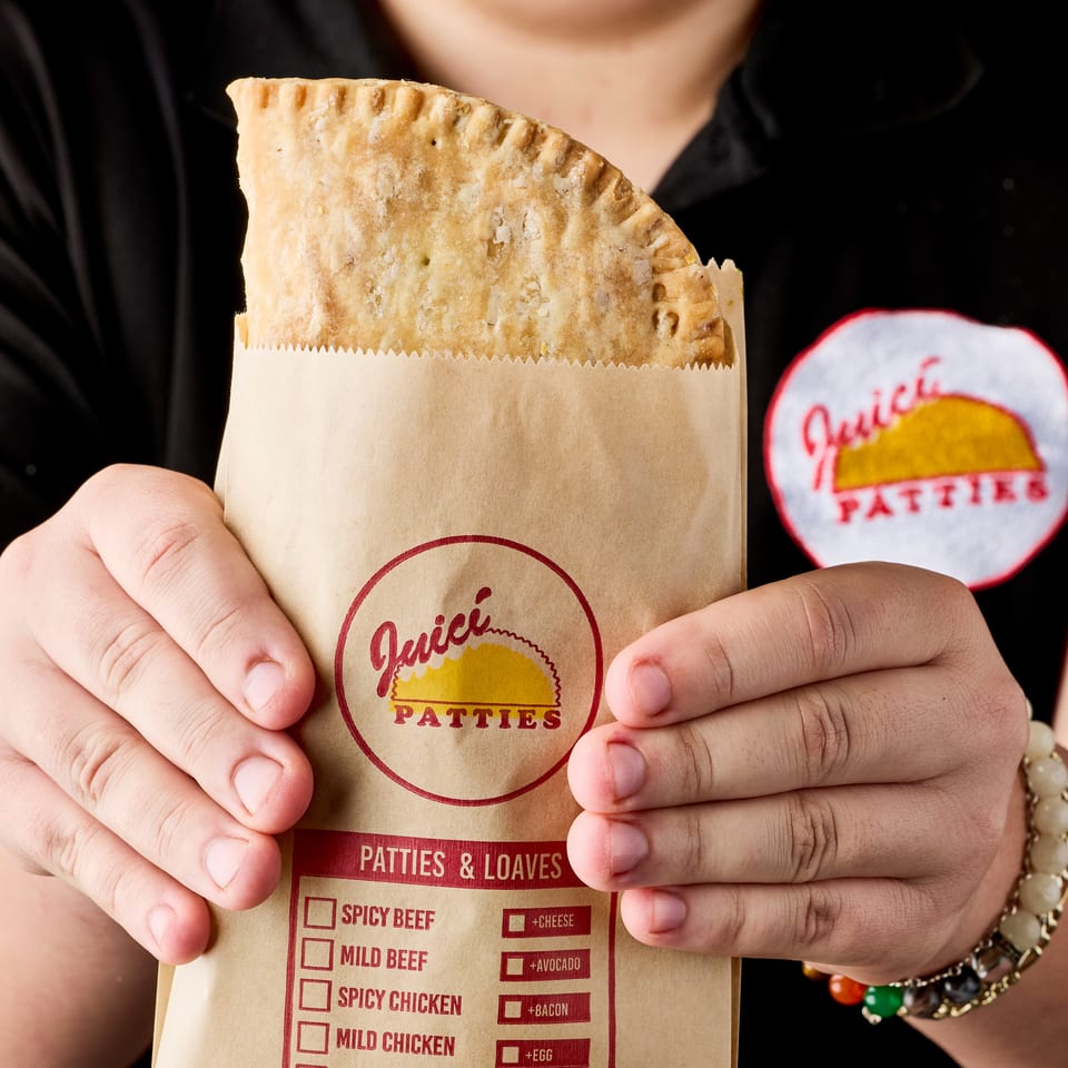 Person holds a golden Jamaican patty in branded paper packaging labeled Patties & Loaves.