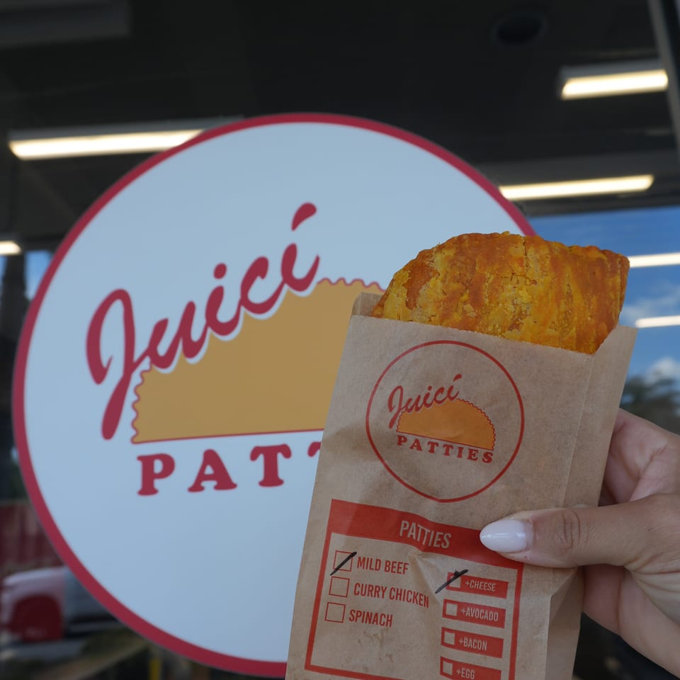 Hand holds a crispy patty in branded paper packaging outside a Jamaican food restaurant.