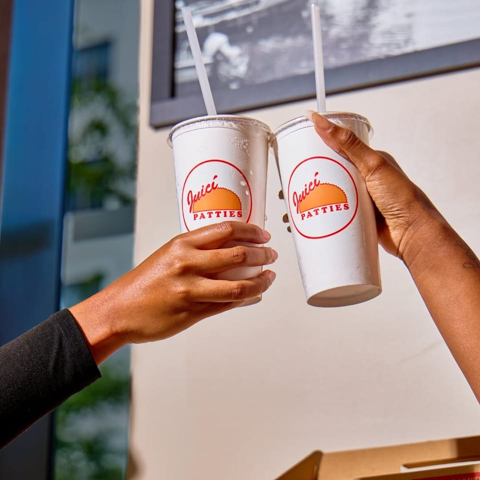 Two hands hold Juicy Patties branded drinks with straws above a takeout box inside a bright restaurant.