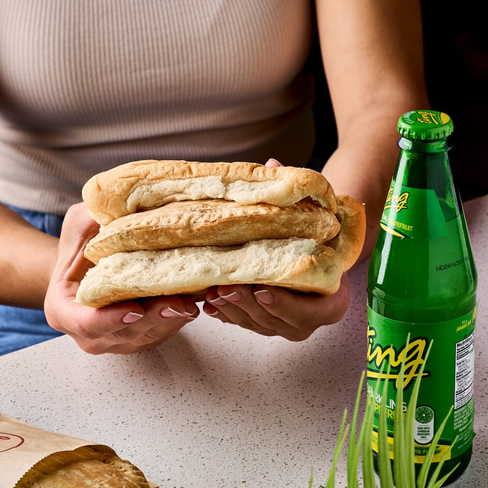 Person holds stacked meat-filled sandwiches beside a green bottle of Ting grapefruit soda on a countertop.