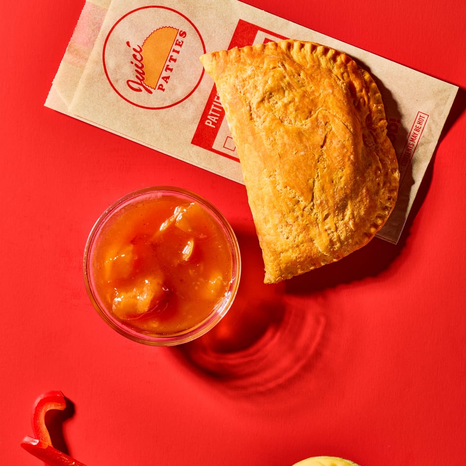Golden empanada on branded paper with orange dipping sauce, sliced red chilies, and mango against a vibrant red background.