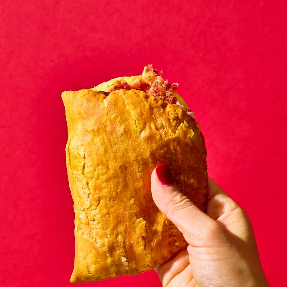 Hand holds a golden-brown triangular stuffed pastry with crispy bacon filling against a vivid red background.