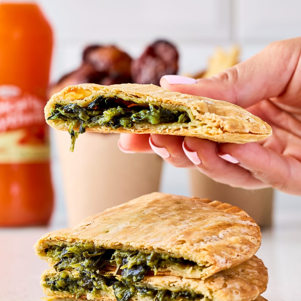 Hand holds a flaky pastry stuffed with leafy greens above a stack of similar savory pastries.