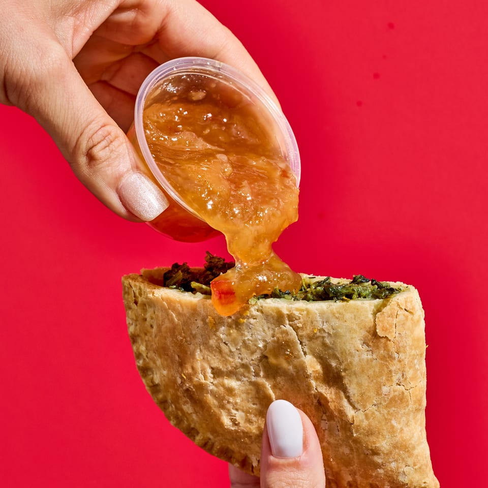 Hand pours thick orange sauce from a cup onto a toasted sandwich filled with greens against a bright red background.