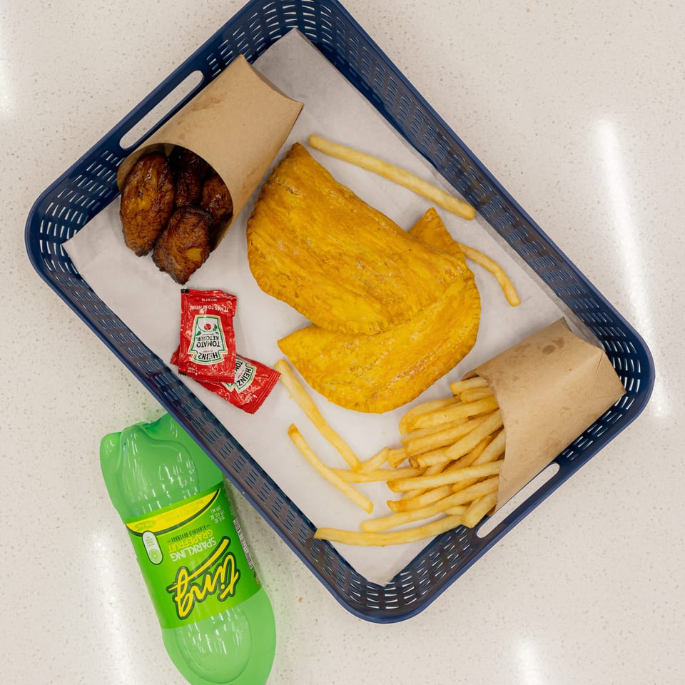 Basket holding golden fried pastries, grilled plantains, French fries, ketchup packets, and a bottle of lime soda.