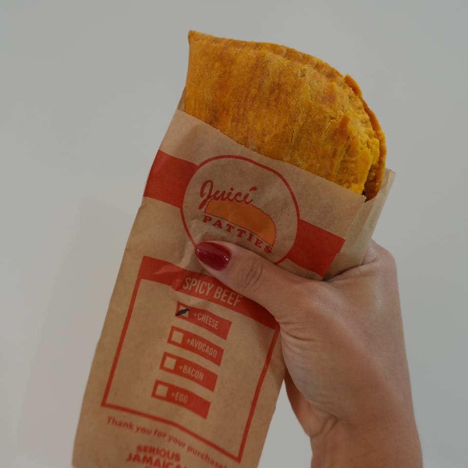 A hand holds a golden Jamaican beef patty in a red-and-tan paper wrapper labeled spicy beef.
