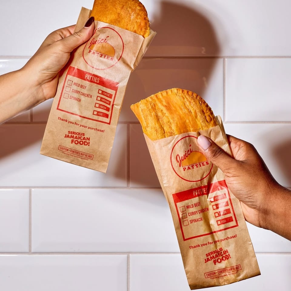 Two hands hold golden Jamaican patties in branded paper sleeves against a white tiled wall.