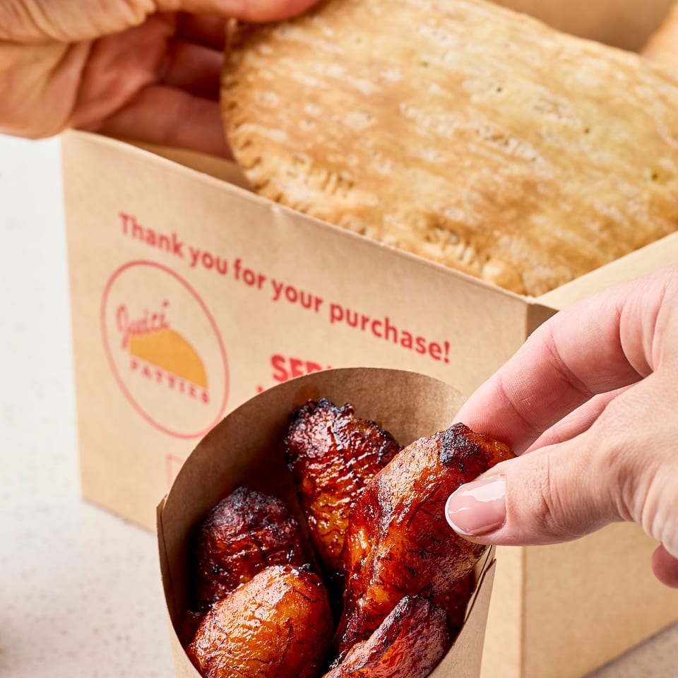 Hand picks up a glazed, grilled chicken wing from a cardboard container beside a box of flatbread.