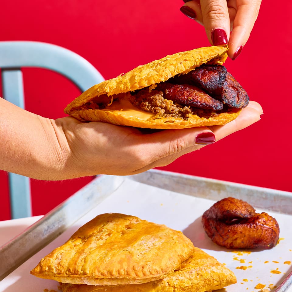 Hands hold an open flaky pastry sandwich filled with grilled meat and minced filling above a tray of golden pastries.