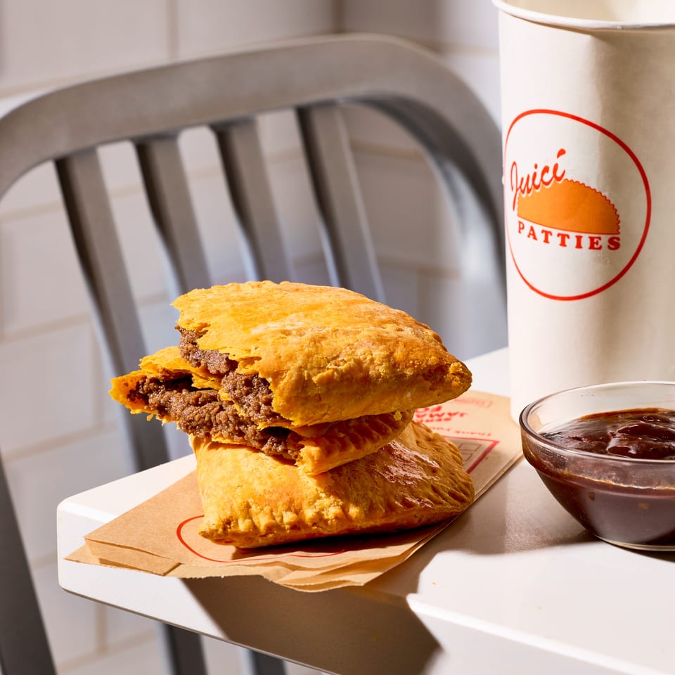 Stacked flaky meat-filled pastries with dipping sauce and a branded drink on a sunlit table.