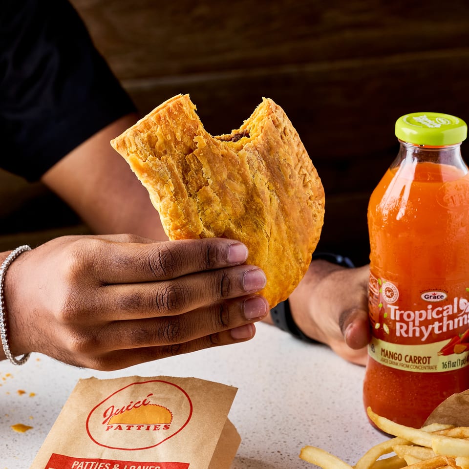Person holds a flaky Jamaican patty beside fries, a Juici Patties bag, and a bottle of Grace Tropical Rhythms drink.