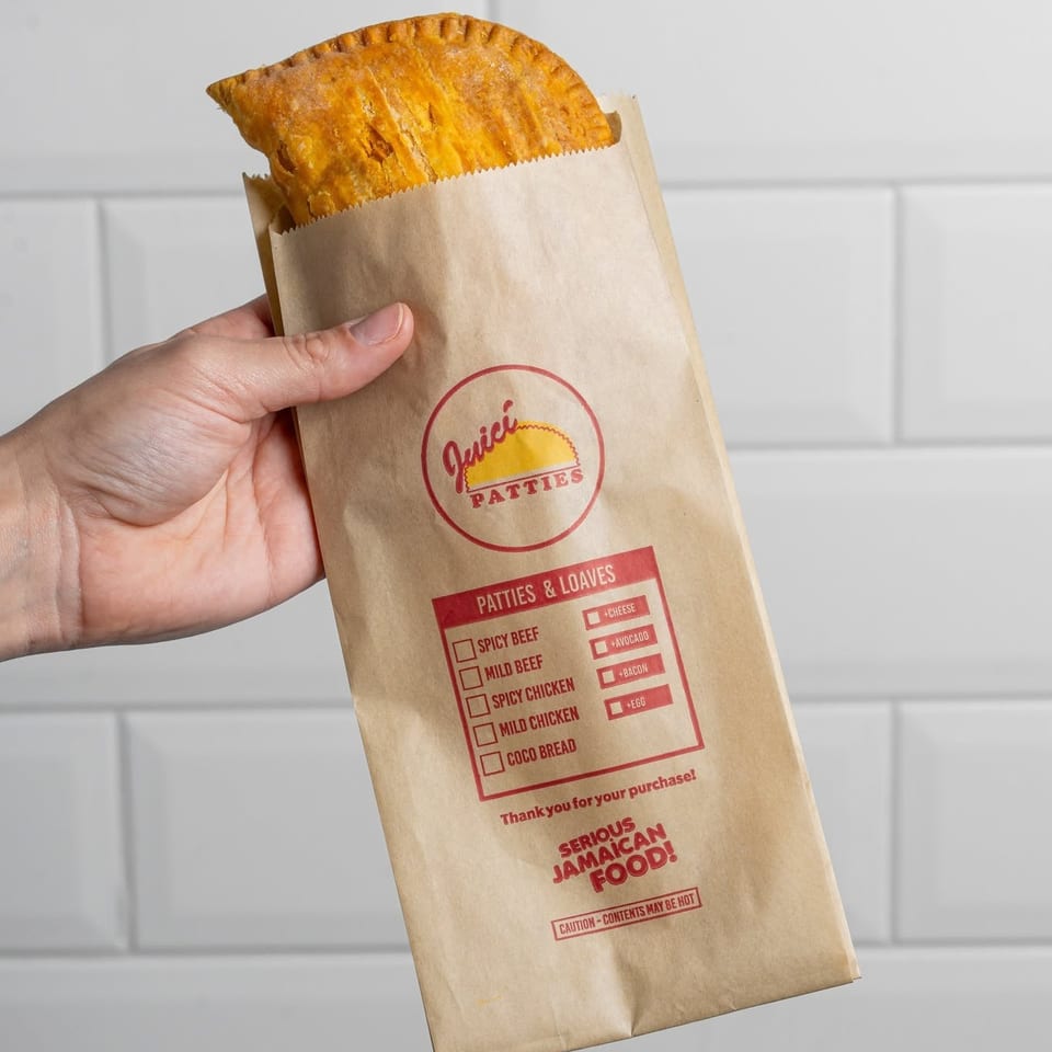 Hand holds a golden Jamaican patty in a branded paper bag against a white tiled wall.