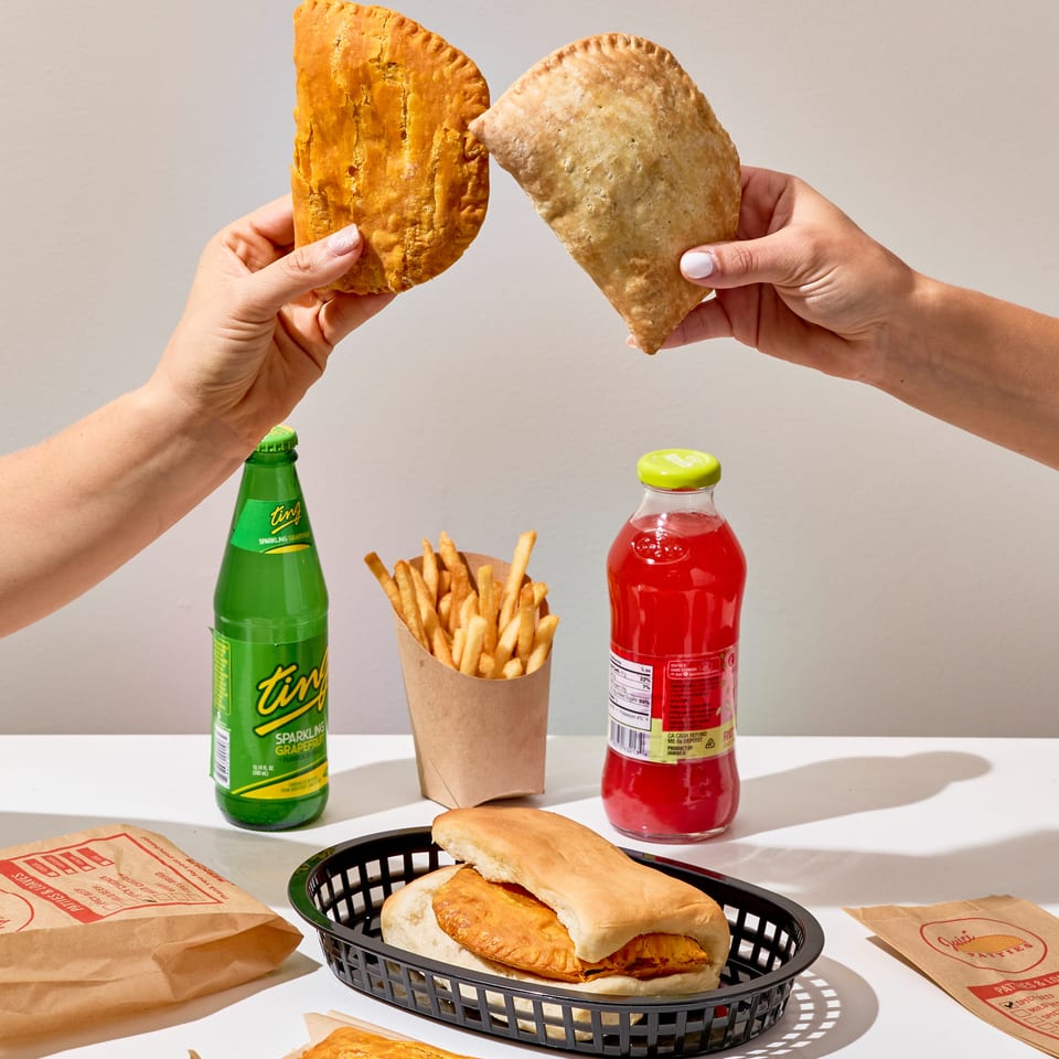 Hands hold two golden Jamaican patties above fries, sandwiches, drinks, and additional patties on a restaurant table.